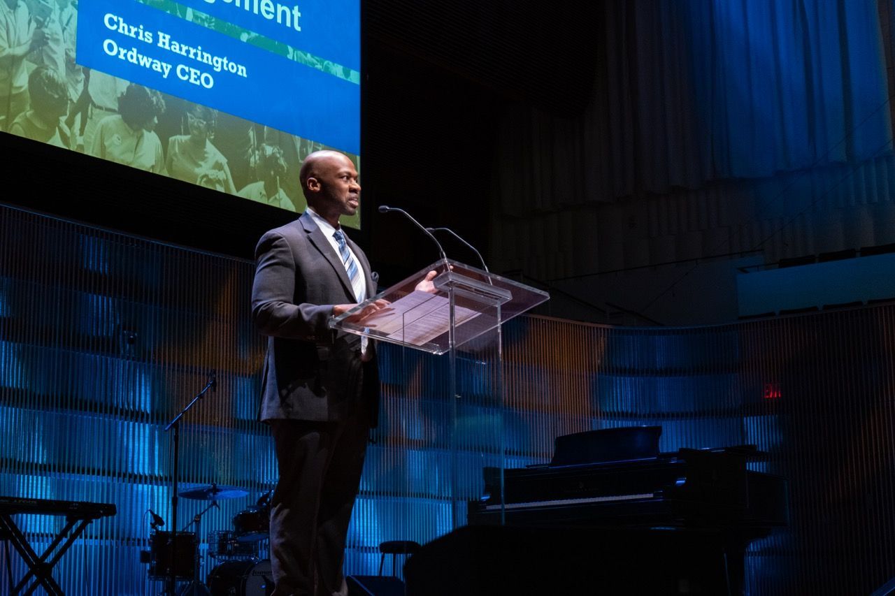 Man in a gray suit speaking at a clear podium on a stage with a blue-lit background, a piano, and musical instruments behind him. A screen above shows the name "Chris Harrington Ordway CEO."